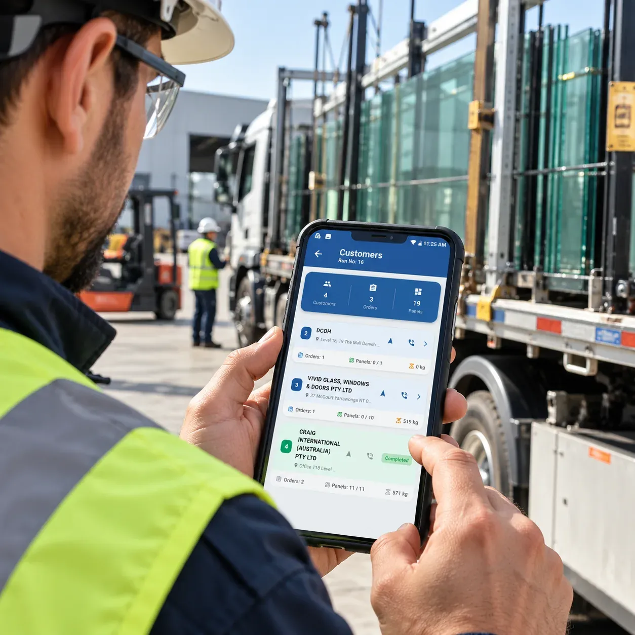 A worker in a hard hat, safety glasses and high-visibility jacket holding a phone with the SPIL Delivery Scanner customer list for a run, beside a truck loaded with glass